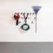 A wall-mounted garage tool organizer with silver hooks holding a red and black chainsaw, a green coiled garden hose, and a blue rake against a white paneled wall.