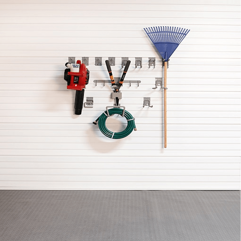 A wall-mounted garage tool organizer with silver hooks holding a red and black chainsaw, a green coiled garden hose, and a blue rake against a white paneled wall.