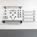 A set of dark gray metal wall-mounted shelves, baskets, and hooks arranged on a white slatwall in a clean indoor setting with a gray floor.