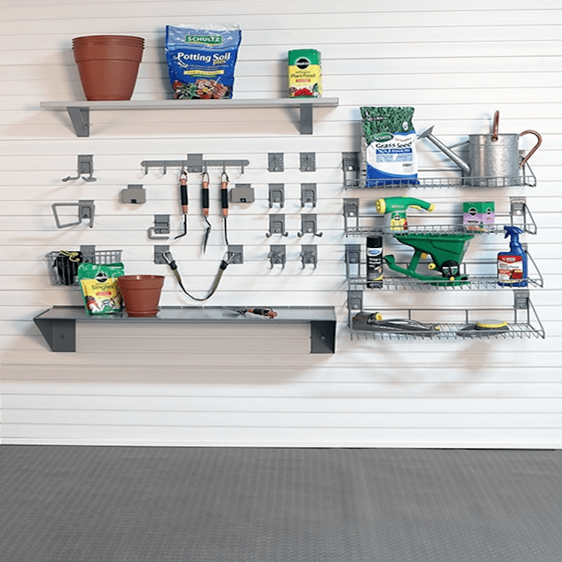 A wall-mounted garage organization system with two gray metal shelves, hooks, and wire racks holding gardening tools and supplies, including a terracotta pot, soil bag, seed bag, watering can, spray bottle, and hand tools, against a white paneled wall and gray garage floor.