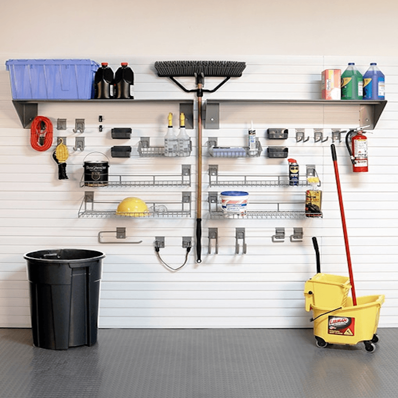 A garage wall organizer setup with a white slatwall, metal shelves, hooks, and various cleaning and storage tools including a broom, mop bucket, trash can, and storage bins.