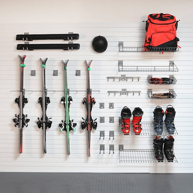 A wall-mounted ski storage system with hooks and wire racks holding four skis, a black helmet, an orange and black backpack, ski boots, and gloves on a white slatted wall.