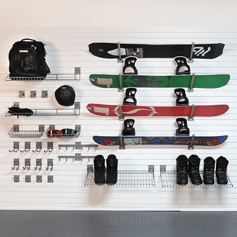 A wall-mounted snowboard storage rack holding multiple snowboards, snow boots, a backpack, a helmet, and goggles on a white slatwall in a garage-like setting.