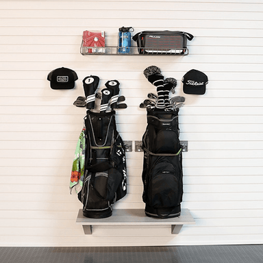 A wall-mounted golf rack with two black golf bags, several golf clubs, two black baseball caps, and a small shelf holding accessories, shown against a white slatwall background.