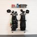 A wall-mounted golf rack with two black golf bags, several golf clubs, two black baseball caps, and a small shelf holding accessories, shown against a white slatwall background.