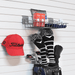 A red Titleist baseball cap hangs on a white slatwall alongside a gray metal wire shelf holding golf accessories, including a red bag, golf balls, striped golf club headcover sleeves, and a golf bag with clubs.
