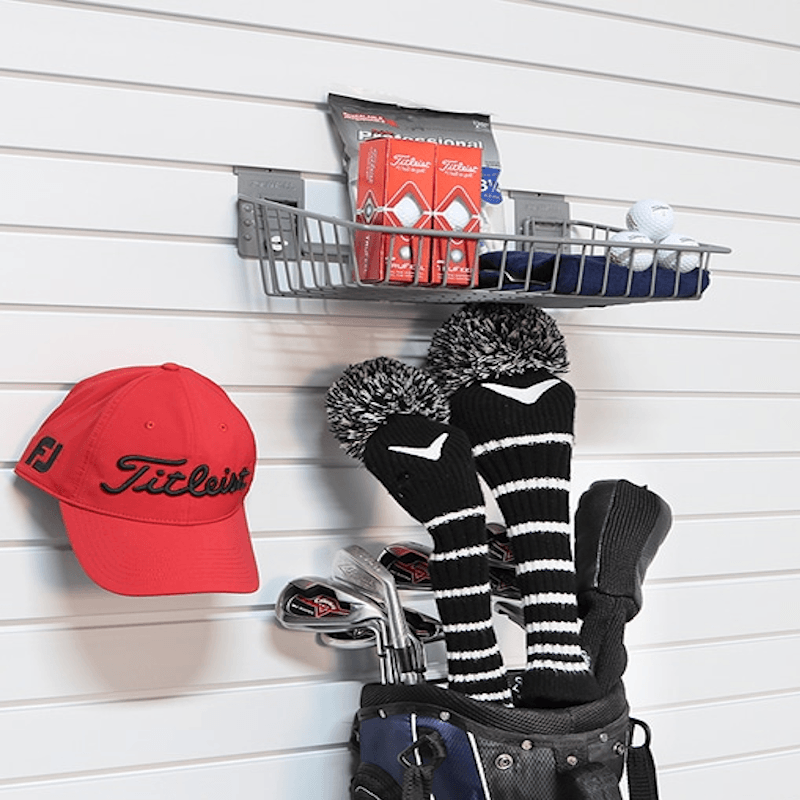 A red Titleist baseball cap hangs on a white slatwall alongside a gray metal wire shelf holding golf accessories, including a red bag, golf balls, striped golf club headcover sleeves, and a golf bag with clubs.