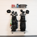 A wall-mounted golf bag rack holding two black golf bags with club heads and tees, plus a top shelf with a water bottle, pouch, and hat, against a white paneled wall.