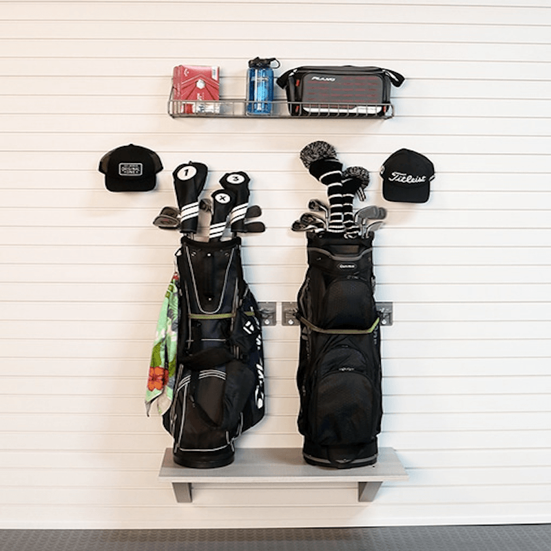 A wall-mounted golf bag rack holding two black golf bags with club heads and tees, plus a top shelf with a water bottle, pouch, and hat, against a white paneled wall.