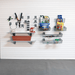 A garage wall organizer setup with white slatwall panels, gray hooks, floating shelves, and wire baskets holding tools, plants, and household items. Visible products include a red planter box, gardening supplies, and packaged goods in the baskets.