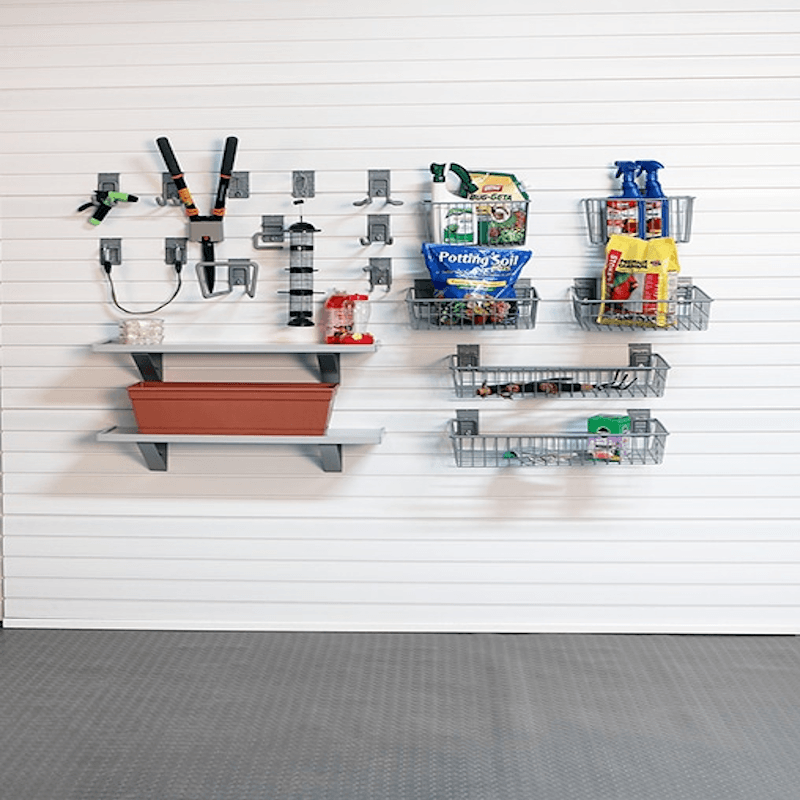 A garage wall organizer setup with white slatwall panels, gray hooks, floating shelves, and wire baskets holding tools, plants, and household items. Visible products include a red planter box, gardening supplies, and packaged goods in the baskets.