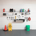 A wall-mounted garage organization system with metal shelves, hooks, and racks holding cleaning supplies, tools, a red fire extinguisher, a yellow warning sign, and a green bucket on a gray garage floor against a white slatwall.