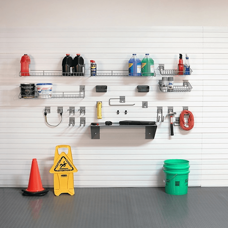 A wall-mounted garage organization system with metal shelves, hooks, and racks holding cleaning supplies, tools, a red fire extinguisher, a yellow warning sign, and a green bucket on a gray garage floor against a white slatwall.