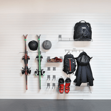 A wall-mounted ski rack system holding skis, helmets, backpacks, a jacket, and ski boots against a white slatted wall.