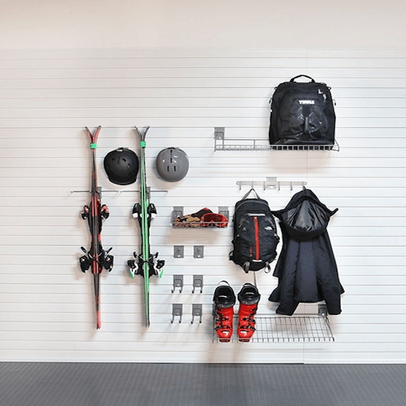 A wall-mounted ski rack system holding skis, helmets, backpacks, a jacket, and ski boots against a white slatted wall.