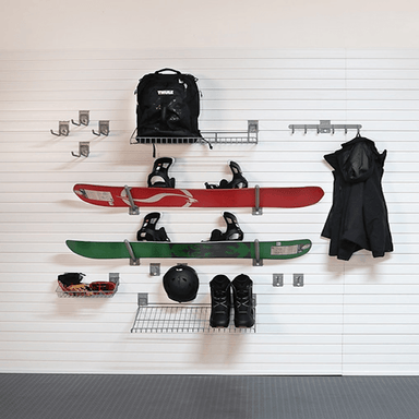 A wall-mounted snowboard rack system holding a red snowboard, a green snowboard, a black backpack, black ski boots, a helmet, sunglasses, and a black coat, against a white slatted wall.