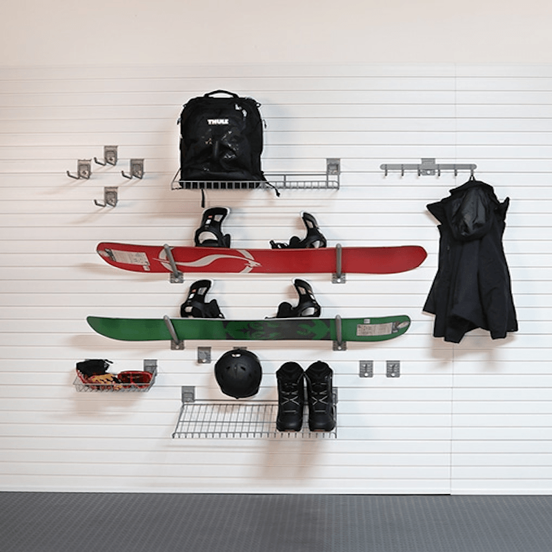 A wall-mounted snowboard rack system holding a red snowboard, a green snowboard, a black backpack, black ski boots, a helmet, sunglasses, and a black coat, against a white slatted wall.