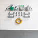A wall-mounted gardening tool organizer set on a white slatwall, including wire shelves, hooks, a hose reel, a green watering can, a metal can, a spray bottle, and a bag of plant fertilizer. A gray textured floor is visible below.