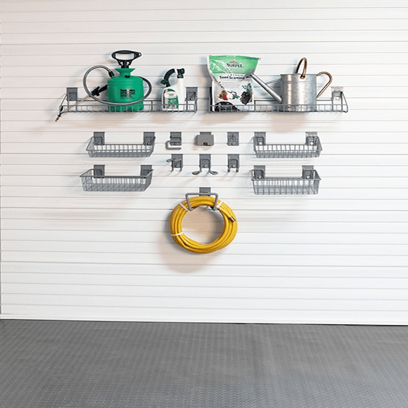 A wall-mounted gardening tool organizer set on a white slatwall, including wire shelves, hooks, a hose reel, a green watering can, a metal can, a spray bottle, and a bag of plant fertilizer. A gray textured floor is visible below.