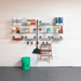 A wall-mounted metal garage organizer rack system with multiple shelves and hooks, shown installed on a white paneled garage wall. The rack holds assorted household cleaning products, a broom, a small wooden step ladder, and a green bucket on the floor.