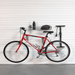 A red mountain bike is mounted on a wall-mounted bike rack system in a garage setting with a white slatted door and gray floor. The rack also holds a black water bottle and a helmet.