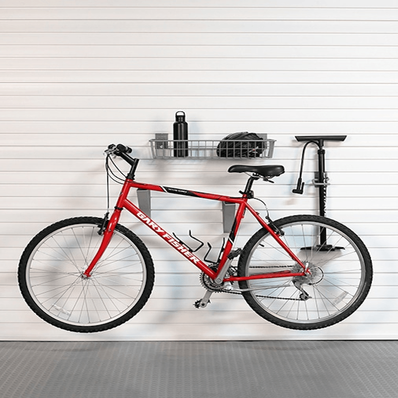 A red mountain bike is mounted on a wall-mounted bike rack system in a garage setting with a white slatted door and gray floor. The rack also holds a black water bottle and a helmet.