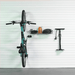 A teal mountain bike is mounted on a white slatted wall in a garage setting, with a black wall-mounted storage basket holding an orange bottle and a black floor pump to the right.