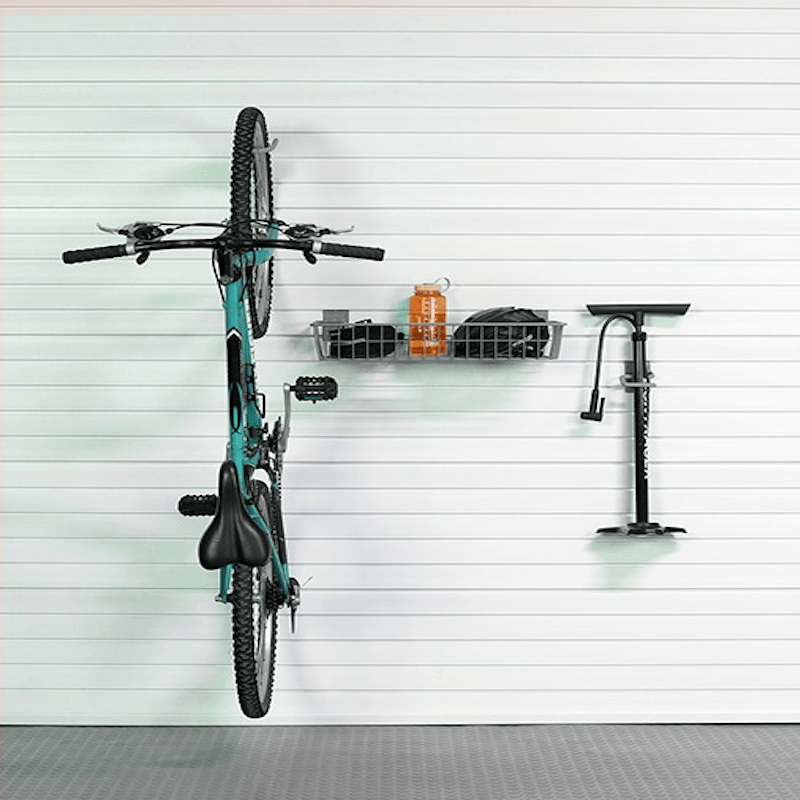 A teal mountain bike is mounted on a white slatted wall in a garage setting, with a black wall-mounted storage basket holding an orange bottle and a black floor pump to the right.
