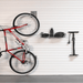 A red mountain bike hangs on a white slatted garage door with wall-mounted storage accessories, including a black basket, a water bottle, a pump, and a small rack. A gray floor mat is visible at the bottom.