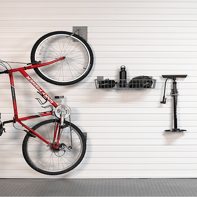 A red mountain bike hangs on a white slatted garage door with wall-mounted storage accessories, including a black basket, a water bottle, a pump, and a small rack. A gray floor mat is visible at the bottom.