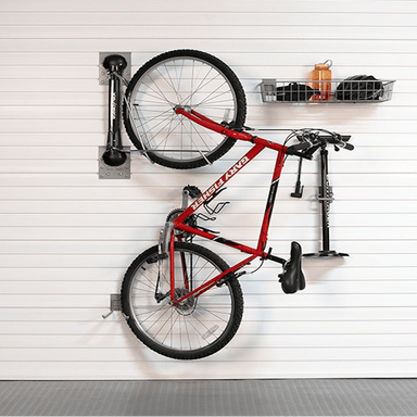 A red bicycle is mounted on a white slatted garage wall using a wall-mounted bike rack system, with a black storage basket and an orange bottle on a shelf to the right.