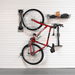A red bicycle is mounted on a white slatted garage wall using a wall-mounted bike rack system, with a black storage basket and an orange bottle on a shelf to the right.