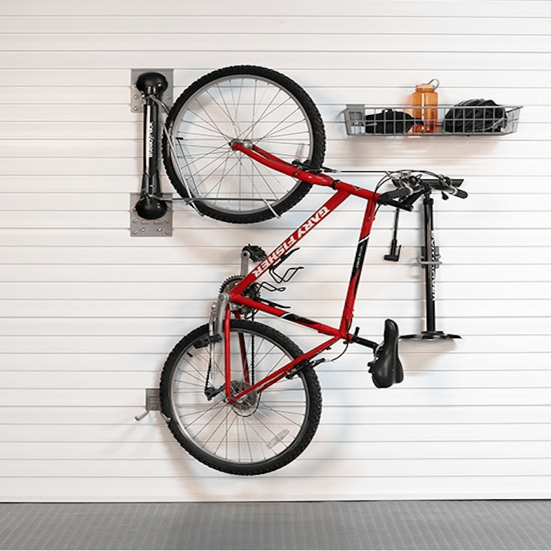 A red bicycle is mounted on a white slatted garage wall using a wall-mounted bike rack system, with a black storage basket and an orange bottle on a shelf to the right.