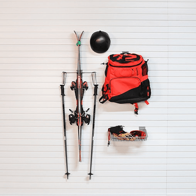 A red and black backpack, ski poles, a black helmet, and ski boots arranged on a white paneled wall.