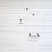 A minimalist set of three dark gray metal wall-mounted storage accessories, including two small square hooks and one wire basket shelf, arranged vertically against a white paneled wall.