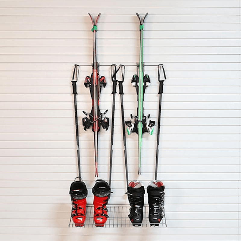 A wall-mounted ski rack holding four skis, four ski poles, and two pairs of ski boots against a white paneled wall.