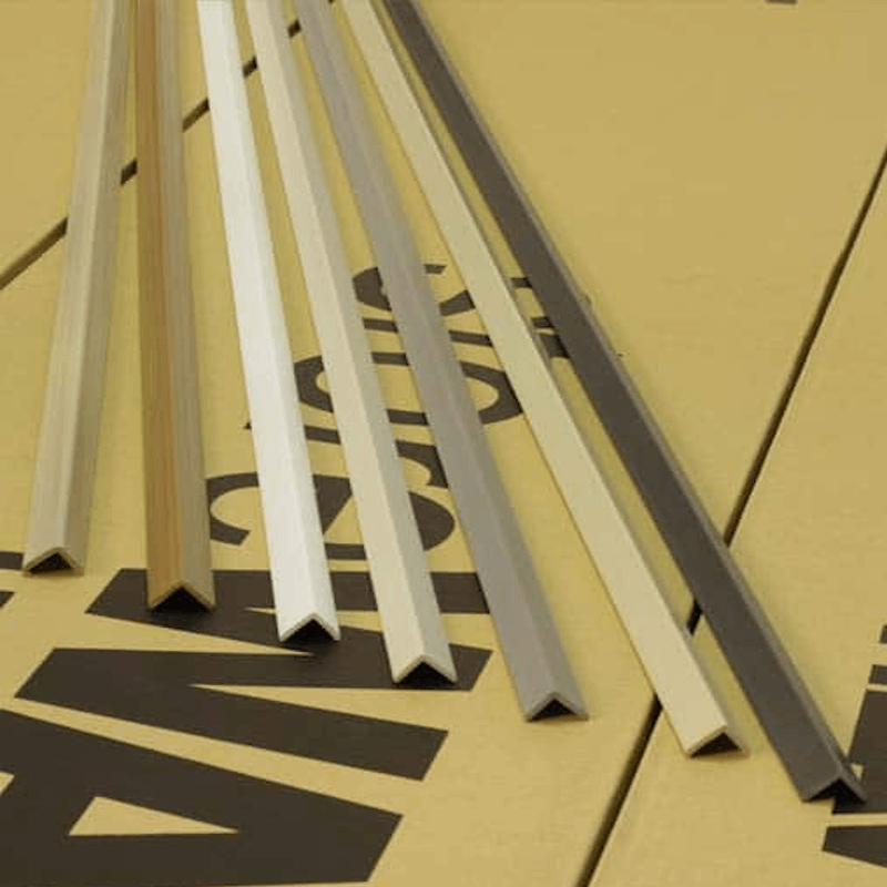 Six aluminum channel profiles in various colors (silver, white, beige, and dark gray) arranged diagonally on a yellow cardboard background with black printed letters.