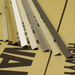 Several slim metal or wood trim profiles shown in different colors, arranged diagonally on a tan surface with bold black printed letters.