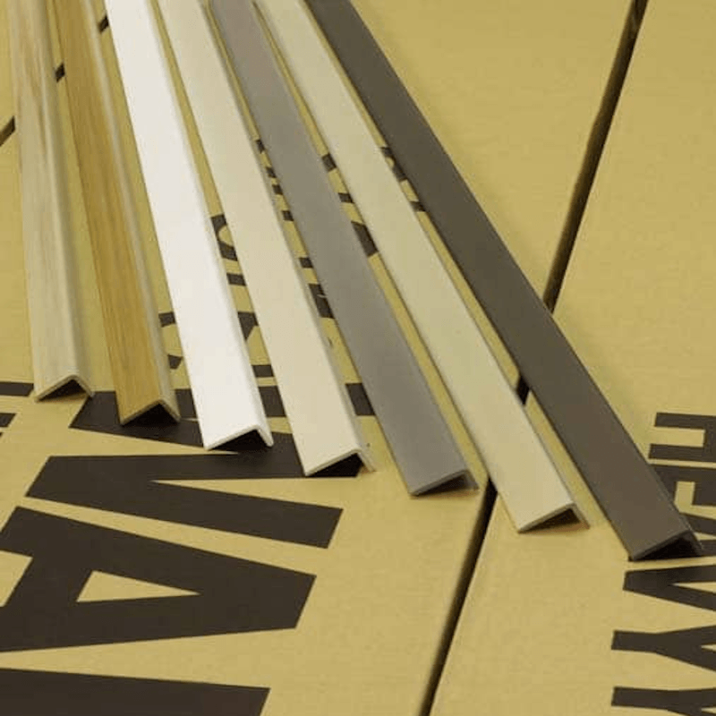 Several slim metal or wood trim profiles shown in different colors, arranged diagonally on a tan surface with bold black printed letters.