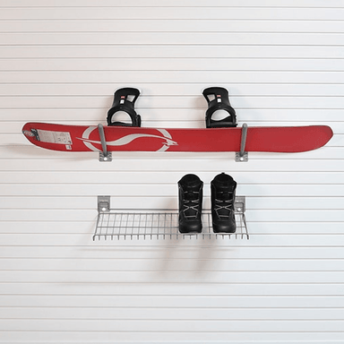 A red snowboard with black bindings mounted on a white slatted wall above a small metal shelf holding a pair of black snow boots.