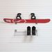 A red snowboard with black bindings mounted on a white slatted wall above a small metal shelf holding a pair of black snow boots.