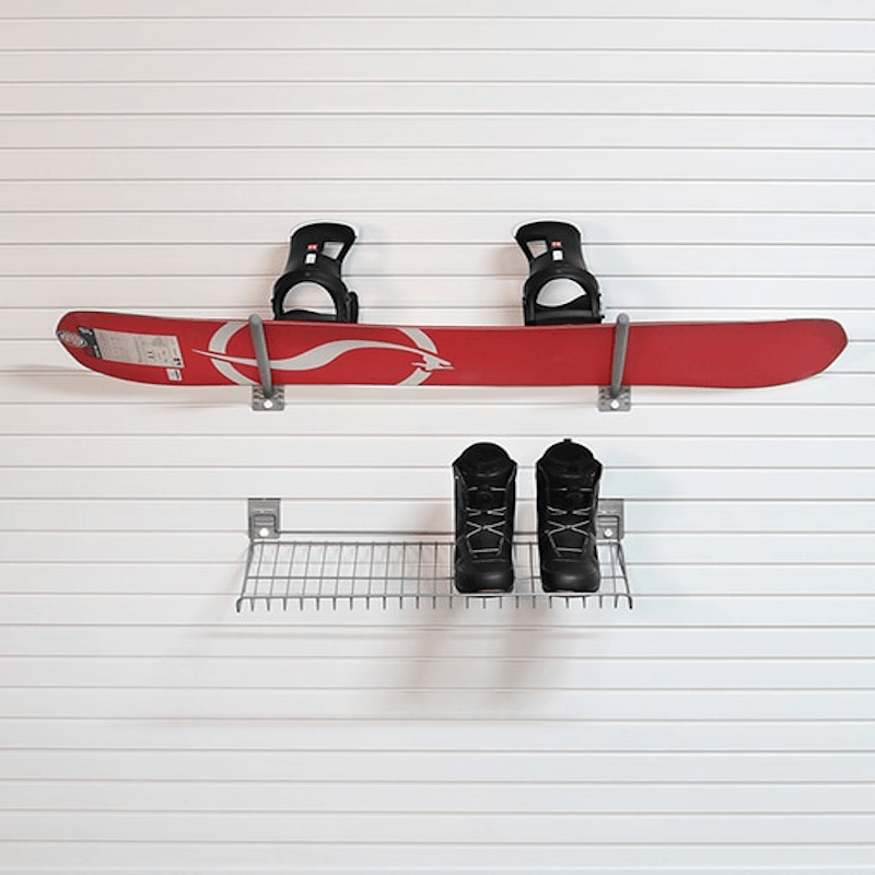 A red snowboard with black bindings mounted on a white slatted wall above a small metal shelf holding a pair of black snow boots.