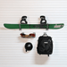 A green snowboard with black bindings mounted on a white slatted wall, with a black helmet, a metal glove rack, and a black Thule backpack hanging below.