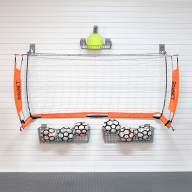 A wall-mounted basketball hoop set with orange side panels labeled BOWNET, a white net, two metal ball racks filled with soccer balls, and a yellow cone on a white paneled wall.