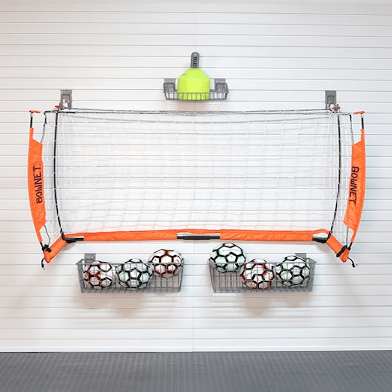 A wall-mounted basketball hoop set with orange side panels labeled BOWNET, a white net, two metal ball racks filled with soccer balls, and a yellow cone on a white paneled wall.