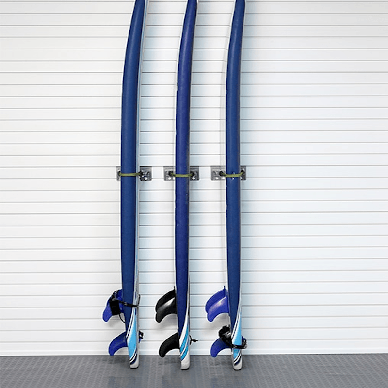 Three blue surfboards or long boards with black and blue fins are mounted on a white slatted wall in a clean indoor setting.