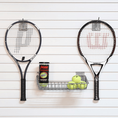 Two tennis rackets mounted on a white slatted wall, with a metal wire basket holding three yellow tennis balls and a can of Denn tennis balls between them.