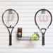 Two tennis rackets mounted on a white slatted wall, with a metal wire basket holding three yellow tennis balls and a can of Denn tennis balls between them.