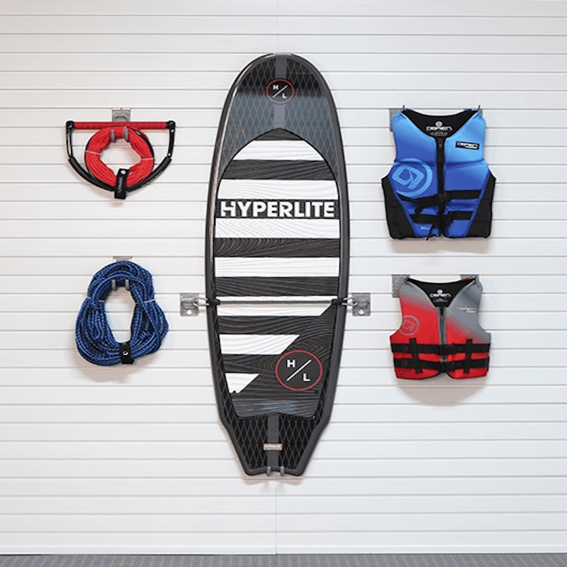 A HYPERLITE wakeboard or ski board is mounted on a white slatted wall, flanked by accessories: a red and black grab handle, a blue rope, and two life vests in blue and red-gray.