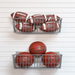 Two gray metal wire baskets mounted on a white slatted wall, filled with sports balls: footballs in the top basket and basketballs in the bottom basket.